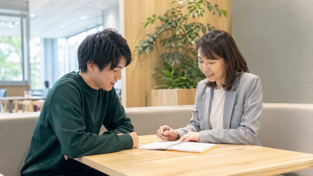 大学生が大学キャリアセンターのような明るい相談スペースで、職員にインターンの相談をしている様子