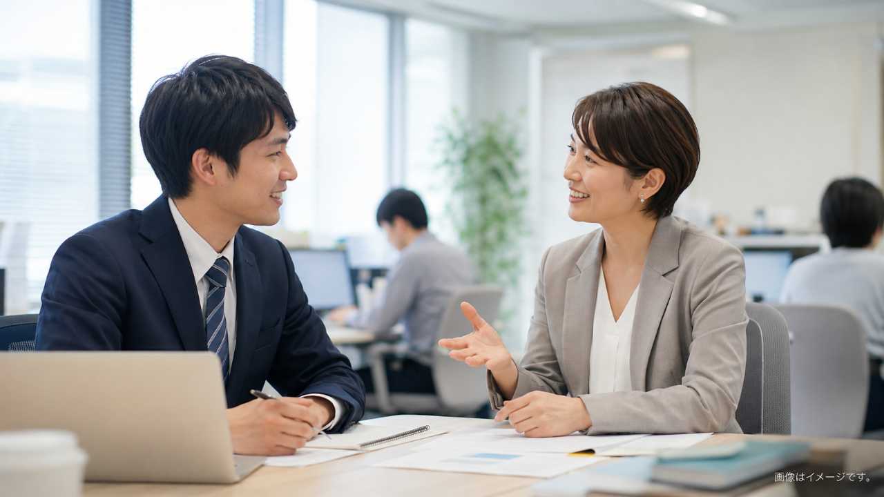 中小企業のオフィスで若手社員が先輩と打ち合わせをしている実写風